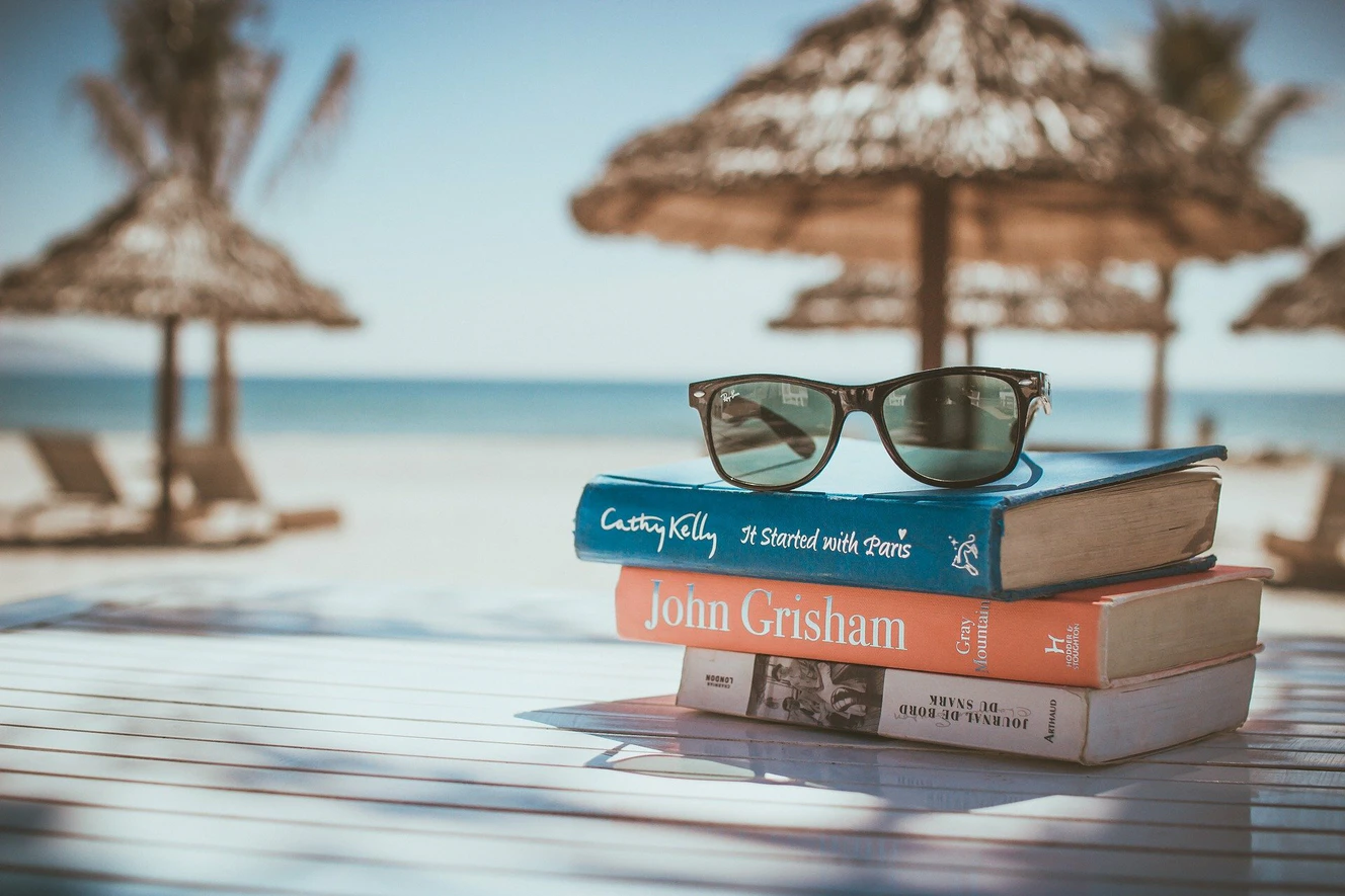 Ein Stapel Bücher am Strand auf denen eine Sonnenbrille liegt